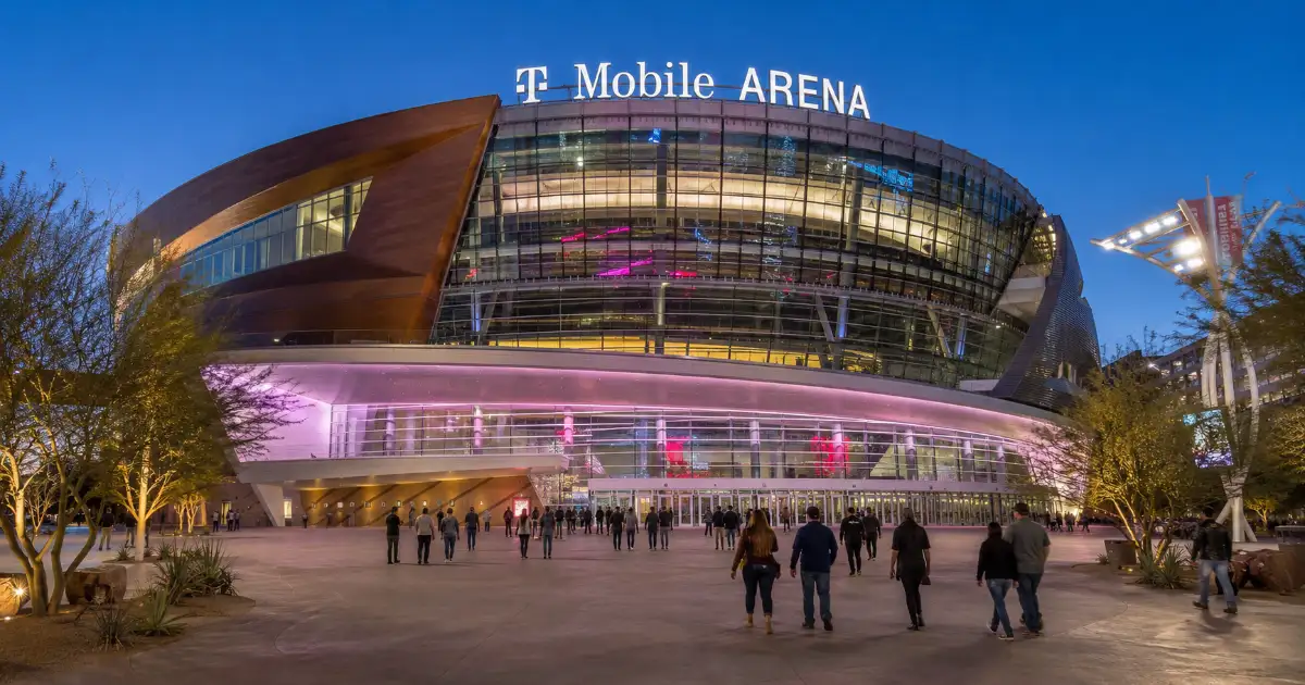 Exterior view of T-Mobile Arena in Las Vegas showing the illuminated glass façade, main entrance, and visitors arriving through Toshiba Plaza.