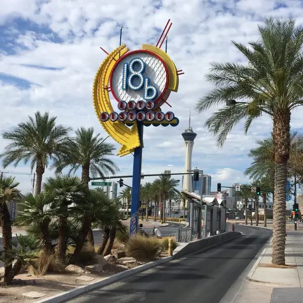 Street view of Las Vegas' 18b Arts District sign with vibrant yellow and red design on a blue pole, set against palm trees, traffic lights, and the iconic Stratosphere Tower—capturing the creative vibe and cultural landmarks of Downtown Las Vegas.