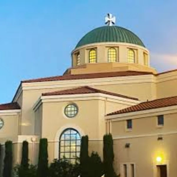 A daytime exterior shot captures the front facade of St. John the Baptist Greek Orthodox Church. The building features a light-colored stone or stucco exterior with distinct Byzantine architectural elements, including rounded arches over the entrance and possibly a dome visible in the background. A prominent, dark-colored cross stands atop the building. The entrance area has a covered walkway or portico supported by columns. Lush green landscaping, including trees and bushes, surrounds the church. A sign bearing the church's name is visible near the entrance. The sky above is bright, suggesting it's daytime. The overall impression is of a serene and established place of worship.