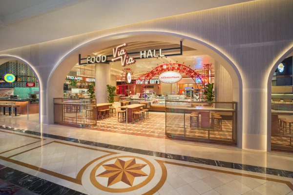 The image shows the entrance to Via Via Food Hall at The Venetian Resort & Casino in Las Vegas. A large, arched sign overhead reads "FOOD HALL" and "Via Via" in bold letters. Just beyond the entrance, the space opens up to a stylish, modern interior with a checkered floor, a variety of vendor signs, and an inviting mix of tables and chairs. Lush greenery adds a touch of warmth, and the space is well-lit, combining natural light with sleek ceiling fixtures. Visible vendor signage includes “Tropicana” and “Hot N Juicy Crawfish,” hinting at the diverse dining options inside. The overall atmosphere is lively and welcoming, blending contemporary design with a hint of old-world charm