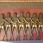 Bronze statue of eight women standing in a row with their backs turned, arms around each other, on display at the original Riviera Hotel & Casino location. Above the statue, a bold sign reads, “Crazy Girls — No ‘IFs’ ‘ANDs’ OR…,” capturing the provocative and iconic spirit of the long-running Vegas revue. The scene reflects classic Las Vegas glamor with a playful edge.