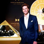 Tom Brady in navy suit stands beside illuminated pyramid display of all seven Super Bowl rings at the Hall of Excellence inside Fontainebleau Las Vegas, celebrating his NFL legacy in a museum curated by Brady and sportscaster Jim Gray.