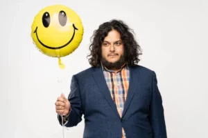 Felipe Esparza, dressed in a blue suit and plaid shirt, holds a yellow smiley-face balloon with a playful grin—blending quirky charm with stand-up swagger against a clean white backdrop.