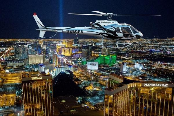 Helicopter flying over Las Vegas Strip at night with Luxor beam and Mandalay Bay skyline in glowing city lights.