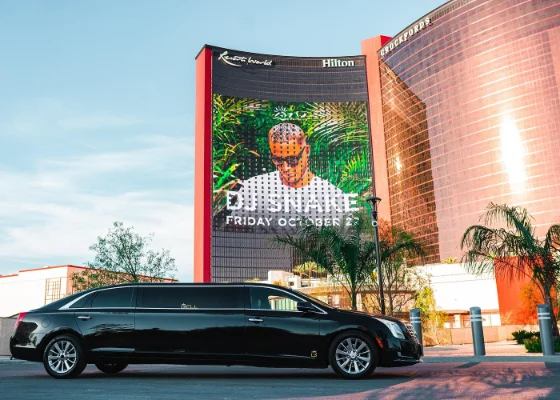 A sleek black limousine is on the Las Vegas Strip providing an airport transfer from Harry Reid Airport to the a hotel. The sky is bright, and palm trees are visible.