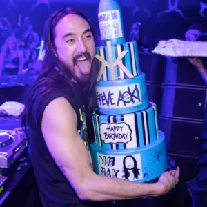 Steve Aoki celebrates his birthday in Las Vegas, holding a multi-tiered blue cake decorated with “HAPPY BIRTHDAY,” surrounded by partygoers and vibrant lighting.