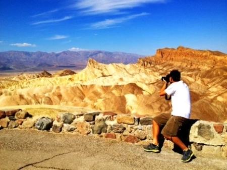 Photographer capturing desert landscape from a scenic viewpoint at Zabriskie Point in Death Valley National Park, with multicolored rock formations, a sharp golden peak, and distant blue mountains under a vivid blue sky.