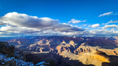 Sweeping winter view of the Grand Canyon with snow-dusted ledges, layered red and orange rock formations, and dramatic lighting beneath a partly cloudy sky, showcasing the canyon’s vastness and seasonal beauty
