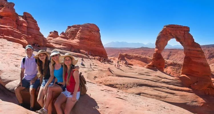 Group of four hikers seated on red sandstone near Delicate Arch in Arches National Park, Utah, with distant mountains and clear blue sky in the background, capturing the scale and scenic beauty of this iconic desert landmark.