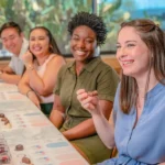 Four guests participate in a guided chocolate tasting at Ethel M Chocolates in Henderson, Nevada. Each person has a curated selection of chocolates and a tasting mat with flavor charts, creating an engaging and educational experience in a bright indoor setting.