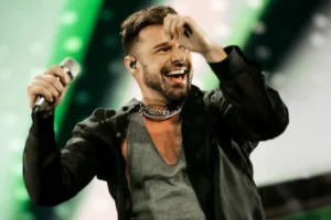 Ricky Martin beams on stage in a sheer black shirt and chain necklace, gripping the mic mid-performance as green and white lights pulse behind him—radiating charisma and Latin pop flair.
