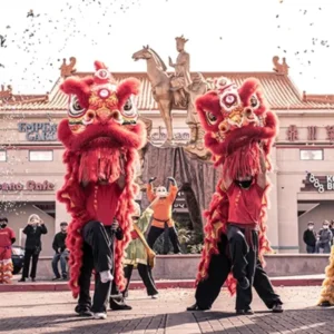 Traditional lion dancers performing during Year of the Dragon celebration at Chinatown Plaza Las Vegas with red and gold costumes, confetti, and festive crowd in front of storefronts and statue