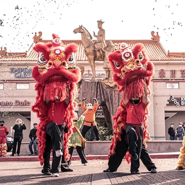 Traditional lion dancers performing during Year of the Dragon celebration at Chinatown Plaza Las Vegas with red and gold costumes, confetti, and festive crowd in front of storefronts and statue