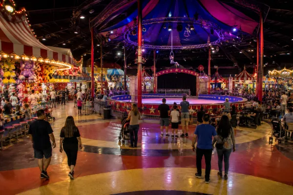 A lively Circus Circus Midway scene with colorful carnival games, prize displays, families, and a distant aerialist above the central ring.