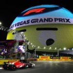 Formula 1 Las Vegas Grand Prix at night with race car speeding past the MSG Sphere displaying digital graphics of a yellow face with eyes moving to watch the cars as they race by