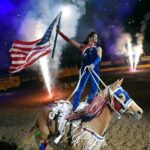 Patriotic rodeo performer in Las Vegas standing on horse with American flag, fireworks, and cheering crowd in arena