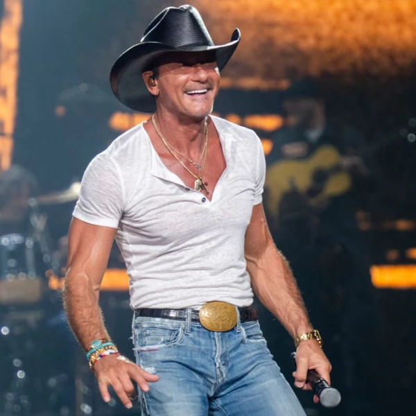 Tim McGraw performing live in black cowboy hat white shirt and jeans with musicians and stage lights in background
