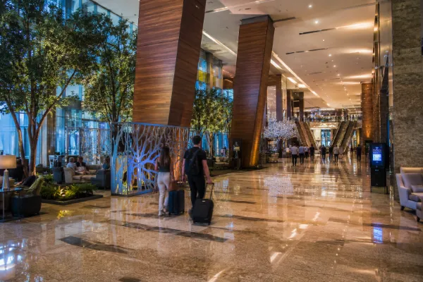 Guests walk through a spacious ARIA interior with polished floors, angled wood columns, indoor trees, and open resort pathways.