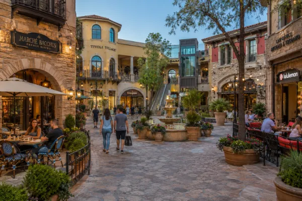Visitors walk among multilevel storefronts, patios, balconies, and landscaped pedestrian paths at Tivoli Village in Las Vegas.