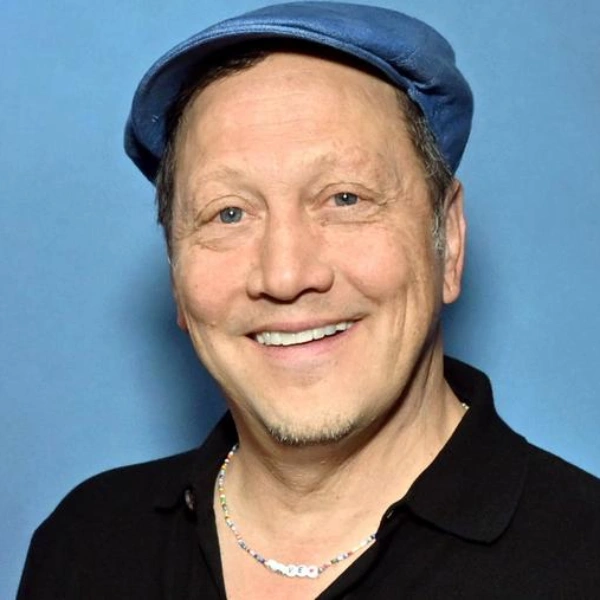 Portrait of comedian Rob Schneider smiling in a blue flat cap and black shirt against a plain blue background