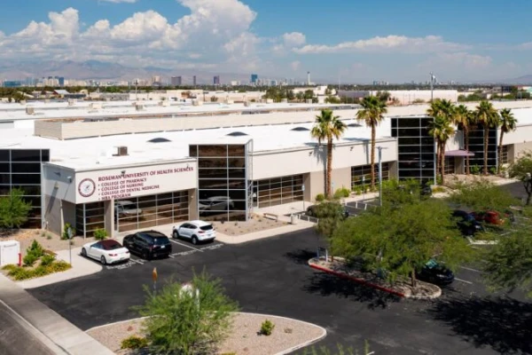 An aerial drone view of the Roseman University of Health Sciences campus in Henderson, Nevada. The modern facility features a white exterior with large glass windows and palm trees in the parking area. The building's signage lists the College of Pharmacy, Nursing, Dental Medicine, and MBA program. The Las Vegas skyline and desert mountains are visible in the distant background under a clear blue sky