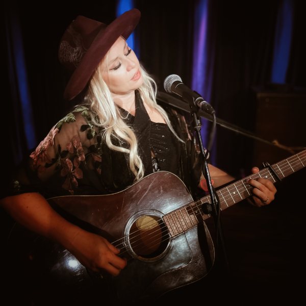 Sofia Talvik performing live onstage with an acoustic guitar, wearing a wide‑brimmed hat and floral-patterned shawl under blue and purple stage lighting