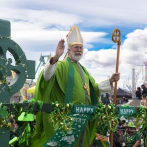 St. Patrick’s Day parade float in Las Vegas featuring a performer in green bishop-style attire holding a staff, surrounded by shamrock decorations with a Ferris wheel and spectators in the background