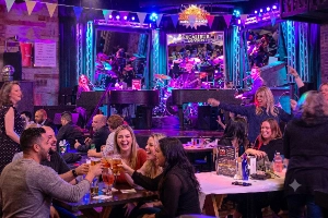 A lively audience enjoying a performance at The Big Bang Dueling Pianos bar at the Excalibur Hotel & Casino in Las Vegas. People in the foreground are laughing, clapping, and toasting beer glasses while two pianists, a drummer, and other musicians play on a blue-and-purple-lit stage.