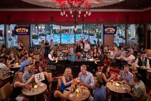 A lively crowd enjoying a live dueling pianos show at the Eastside Lounge in Las Vegas, featuring two pianists entertaining guests who are cheering and toasting drinks, with a brightly lit resort pool visible through the large windows in the background.