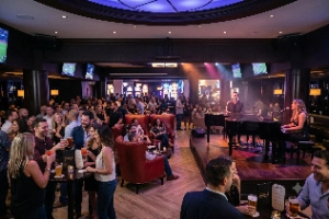 A photograph of a packed dueling pianos show in a dimly lit casino lounge. A female and a male pianist are playing separate grand pianos on a stage under colored spotlights, while a male vocalist sings into a microphone. The audience is crowded around high-top tables and large red leather chairs, talking and drinking. Numerous TVs on the walls display sports games.