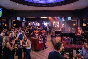 A lively crowd enjoying a dueling pianos show at the Victorian-inspired Juliet Cocktail Room inside The Venetian Resort in Las Vegas. Two pianists are seated back-to-back playing black baby grand pianos while patrons laugh, sing along, and drink signature cocktails in the dimly lit, opulent lounge.