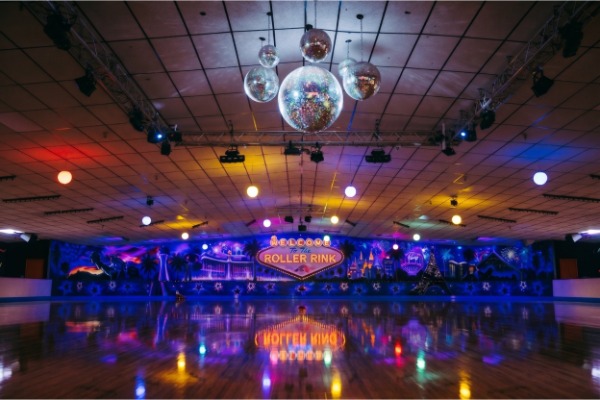 Vibrant indoor roller skating rink filled with local families and skaters cruising the polished hardwood floor under colorful neon lights and glowing disco balls.