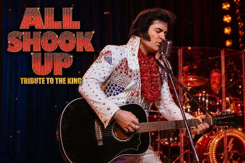 All Shook Up tribute performer in a white rhinestone jumpsuit sings with a black guitar under red marquee lights in Las Vegas.