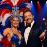Blue-sequined showgirl in a red, white, and blue feathered headdress stands with a tuxedoed host for America The Show.