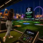 Guest hitting golf balls from a high-tech driving range bay at Atomic Golf Las Vegas with illuminated targets and the Las Vegas Strip skyline in the background.