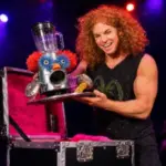 Carrot Top smiles while holding an absurd homemade comedy prop under colorful stage lights.
