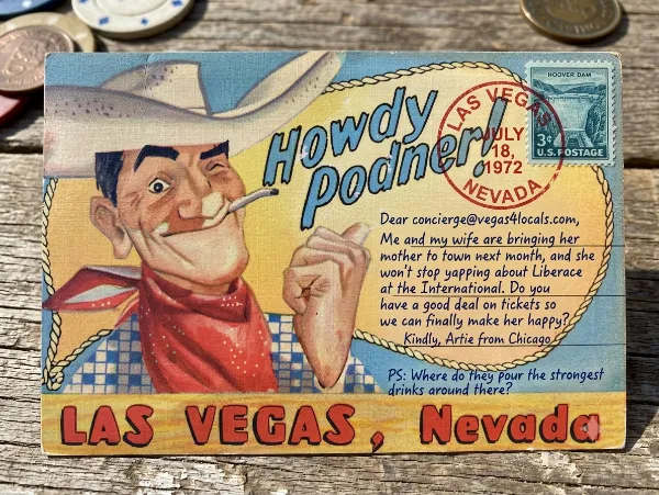 A vintage 1970s Las Vegas postcard featuring a winking cowboy. The message area contains a handwritten cursive inquiry dated July 18, 1972, addressed to concierge@vegas4locals.com. Artie from Chicago asks for ticket deals to see Liberace at the International Hotel to please his mother-in-law, and adds a postscript asking where to find the strongest drinks in town. It includes a blue Hoover Dam stamp and a red Las Vegas postmark.