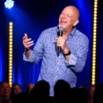 Comedian Don Barnhart laughs with a microphone beneath blue and gold stage lights at Delirious Comedy Club.
