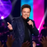 Donny Osmond smiles with a microphone against colorful blue, purple, and magenta Las Vegas stage lighting.