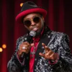Eddie Griffin performs with a microphone while wearing his distinctive red-brimmed hat and sparkling stage jacket.