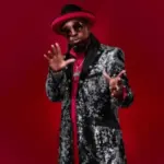Eddie Griffin poses onstage in a long black coat, wearing a black hat with a red rim.