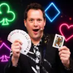 Comedy magician Farrell Dillon displays a fan of playing cards against a colorful neon stage background.
