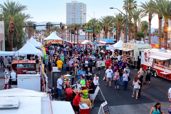A vibrant aerial view of the First Friday street festival in downtown Las Vegas. The photo shows the Arts District's tree-lined street packed with a large crowd of diverse attendees, numerous food trucks, and white canopies under a clear sky, with a tall apartment building in the distance.
