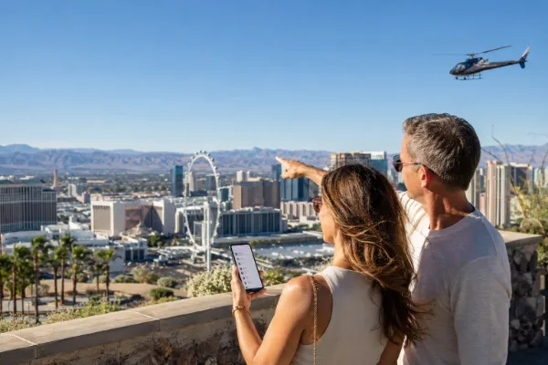 Couple planning Las Vegas tours and attractions from a scenic overlook with the High Roller, Strip skyline, and a sightseeing helicopter in view.