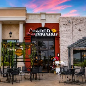 Exterior entrance and front patio of Loaded Empanadas in Boca Park, Las Vegas, with outdoor seating beneath a colorful pink sunset sky.