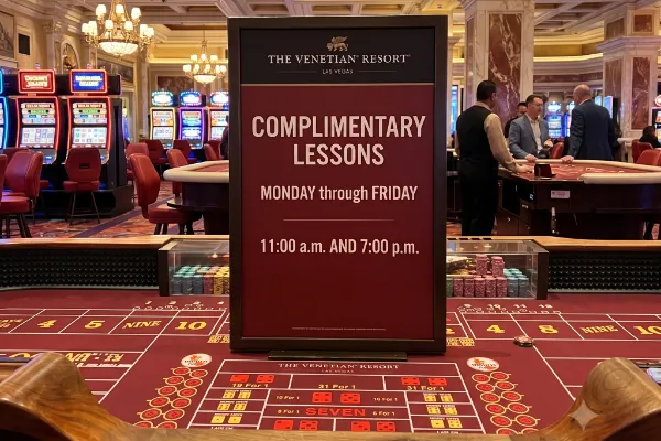 A photo from behind a craps table at The Venetian Resort Las Vegas, showing a large central sign promoting complimentary gambling lessons on weekdays. The sign features the Venetian logo and details times. The table has red felt with betting lines and stacks of chips, and the ornate casino ceiling with frescoes is visible in the background.