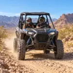 Two helmeted riders securely harnessed inside an RZR on a rocky Mojave Desert trail.