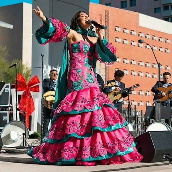 Singer in a pink and teal folkloric dress performing outdoors with mariachi musicians.