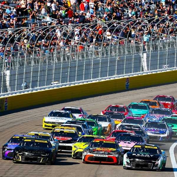 NASCAR race cars competing in a pack beside the catch fence and crowded grandstands.