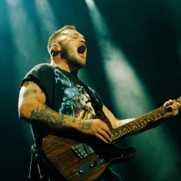 Zach Bryan singing and playing an electric guitar under green stage lights.